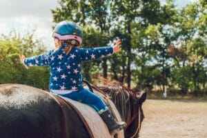 A child confidently rides a horse outdoors, wearing a helmet and star-patterned jacket.
